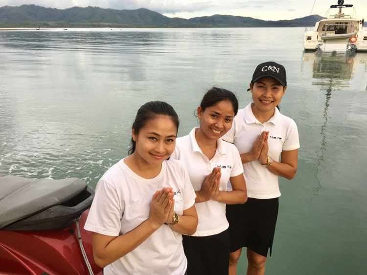 Three Thai women and namaste wishers by a boat on the water welcome guests to Thailand